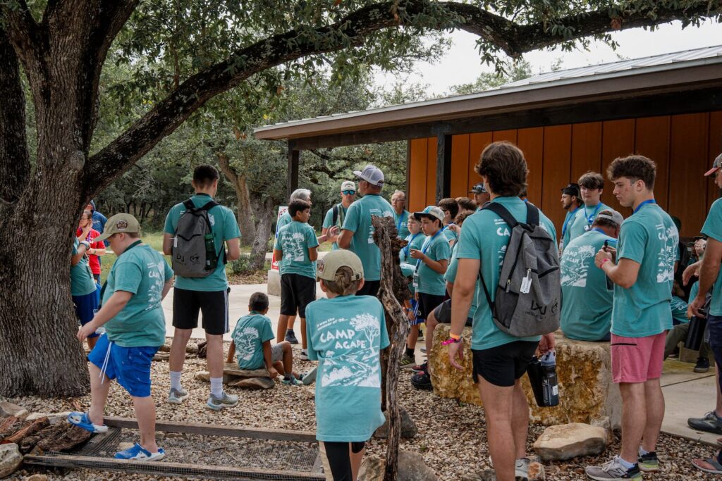 Group of campers in teal Camp Agape shirts gathered outside a wooden pavilion under a large tree, listening to a speaker.