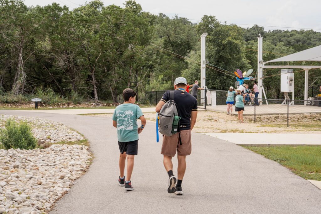 Two youths walk along a paved path toward an outdoor adventure area with a fenced activity station and colorful equipment in the background.