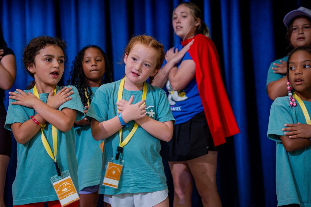Group of children on stage wearing teal shirts and yellow lanyards, performing in front of a blue curtain.