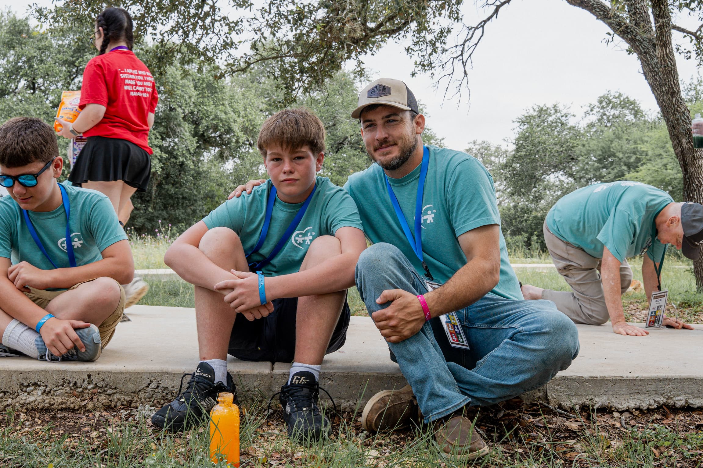 Group of volunteers kneeling outdoors, wearing teal shirts with lanyards, with an orange water bottle in the foreground and trees in the background.