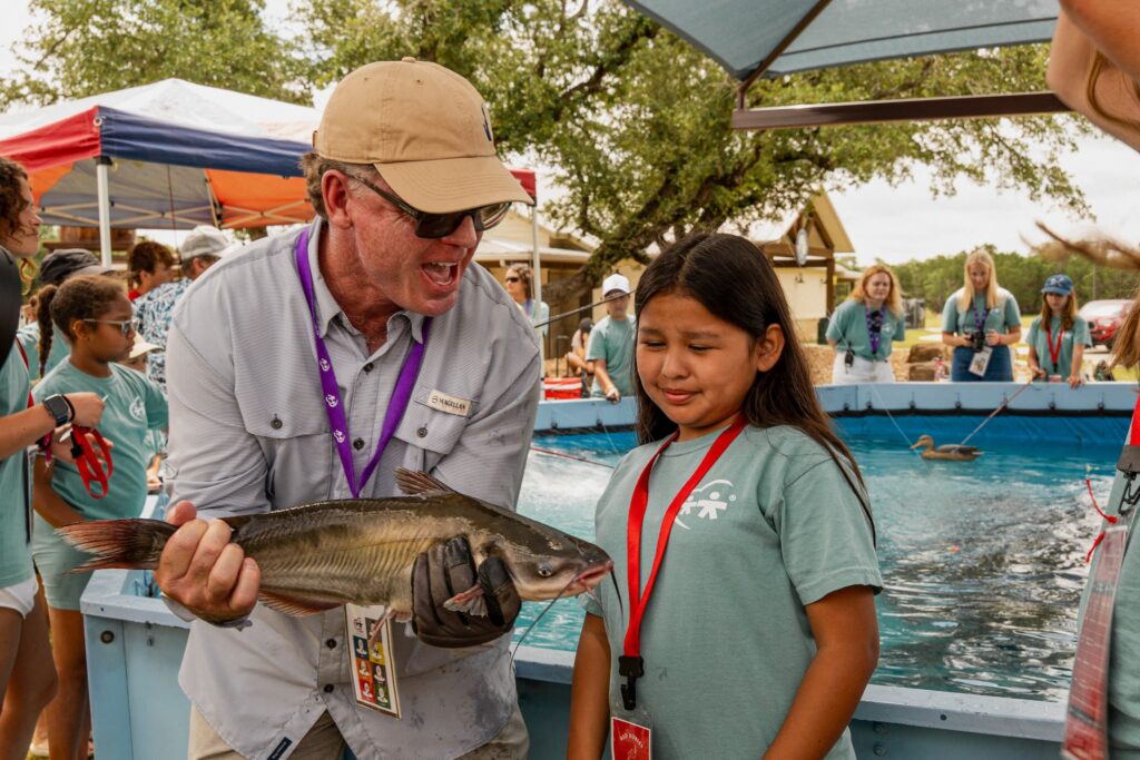 Man in a beige cap and sunglasses proudly holds a large fish for a young girl at a community fishing event by a pool.