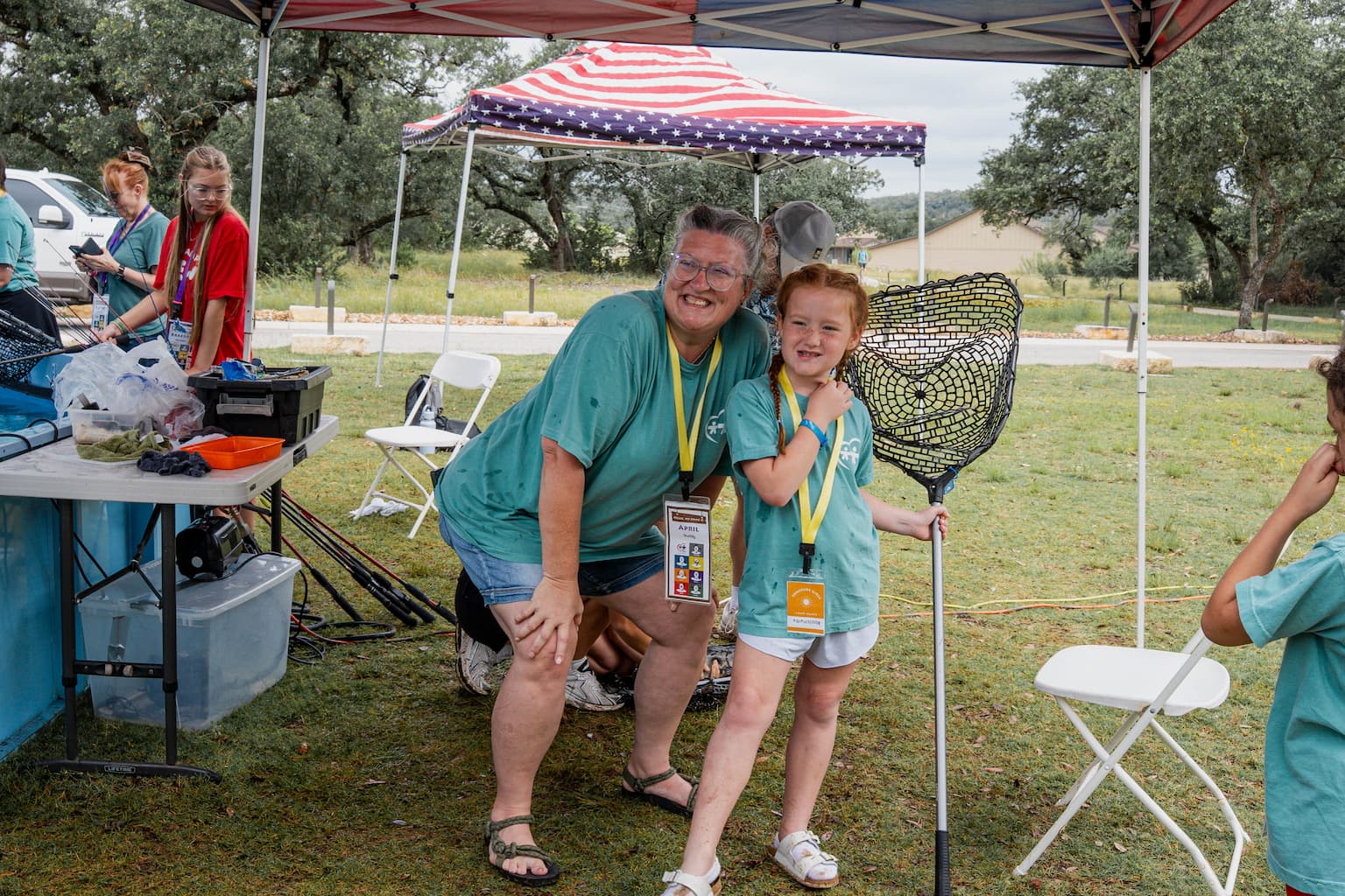 Two volunteers pose with a young camper under a canopy at an outdoor event, both wearing teal shirts and yellow lanyards.
