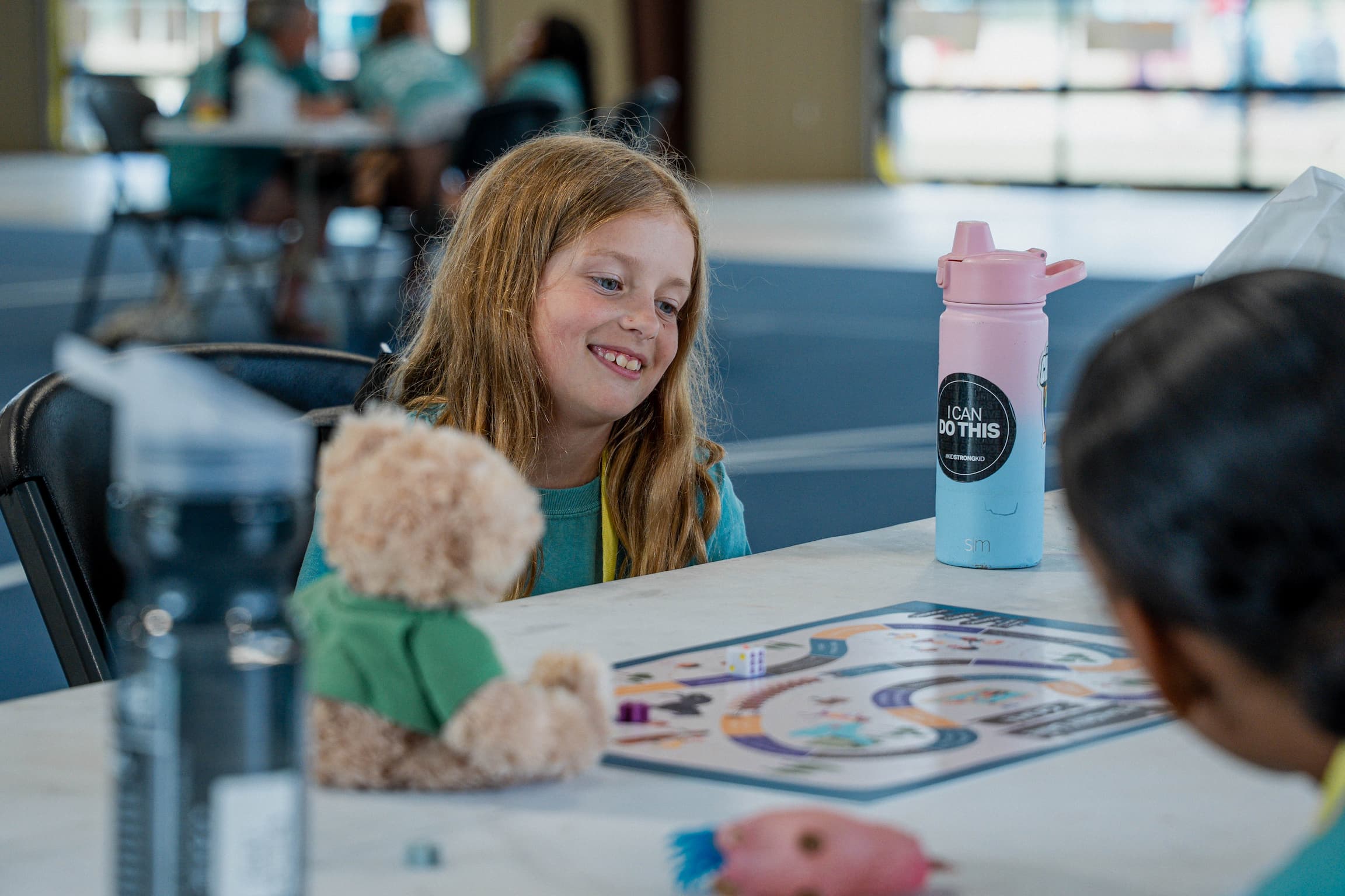 Smiling girl with long hair sits at a table, looking at a colorful board game, with a pink and blue water bottle beside her.