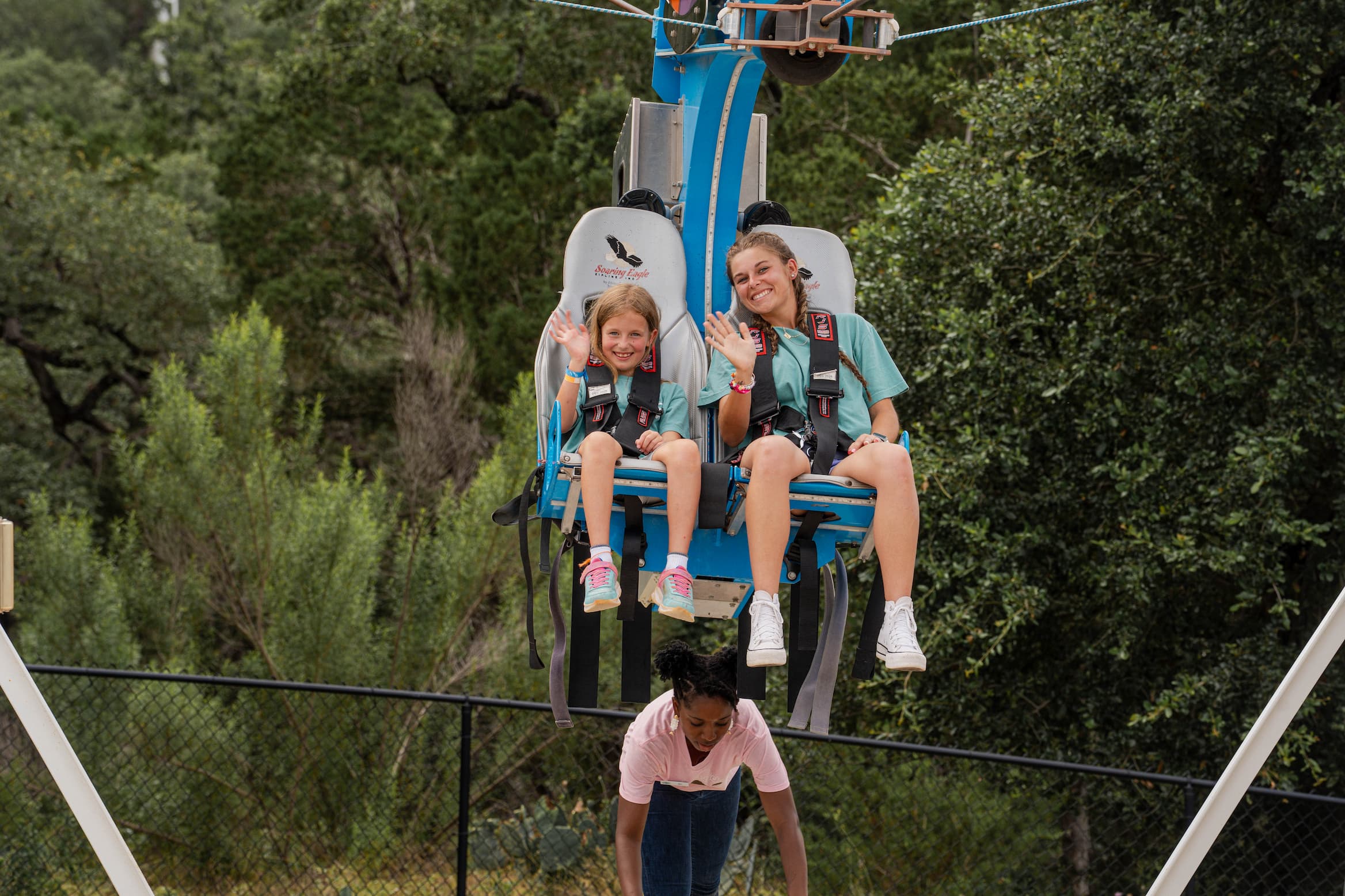 Two smiling girls in teal shirts sit in a harness ride car, waving as they ride above the ground with a staff member nearby.