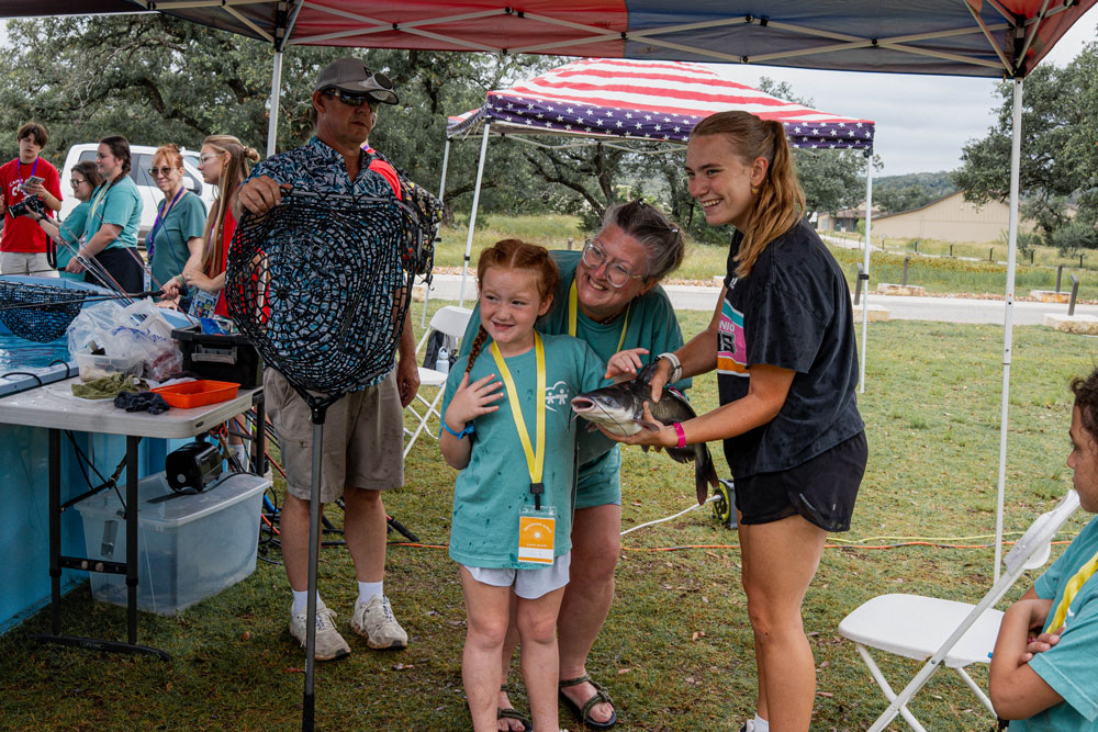 Girl in a teal shirt proudly holds a fish as a volunteer helps her during an outdoor fishing event under a canopy; others watch nearby.