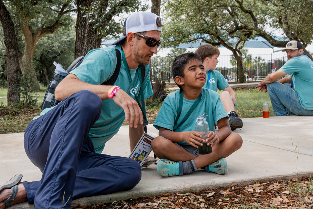 Man in a teal shirt and cap talks with a smiling boy in a matching shirt, both seated on a concrete curb with a jar of drink nearby at an outdoor park.