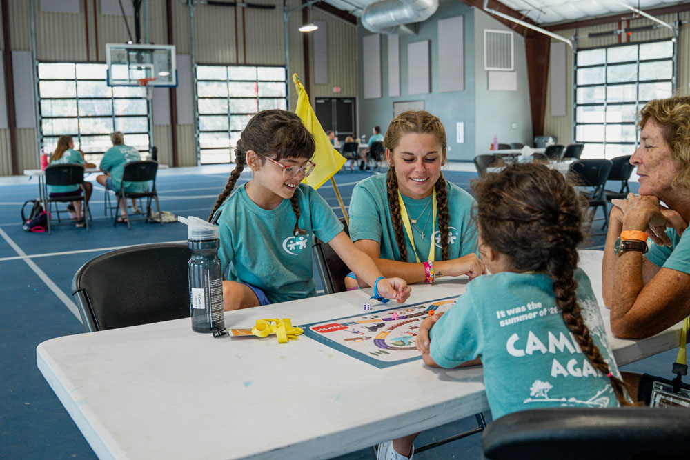 Camper girls and an adult sit at a table in a gym, playing a board game; teal shirts and a water bottle nearby.
