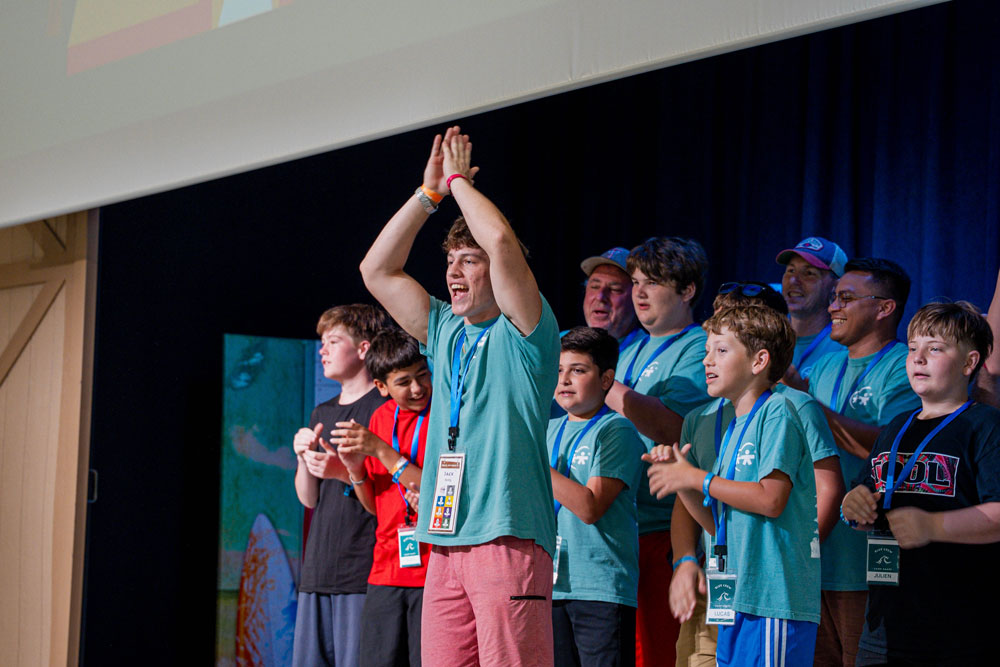Group of young people on a stage celebrating, hands raised in the air, wearing teal T-shirts and lanyards.