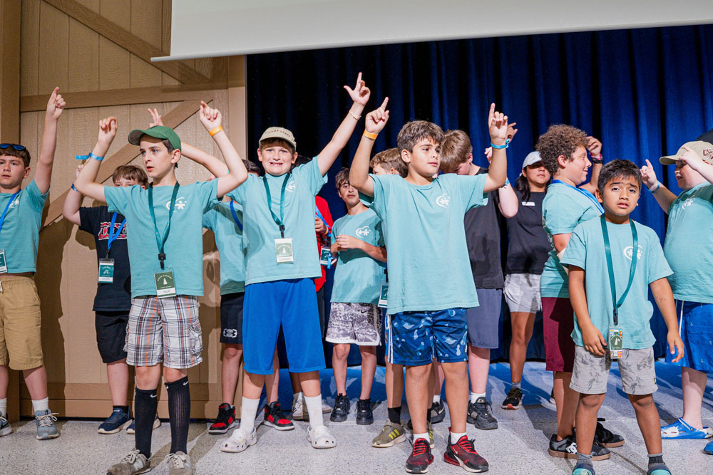 Group of kids in teal shirts on a stage, many with arms raised and wearing lanyards or badges.
