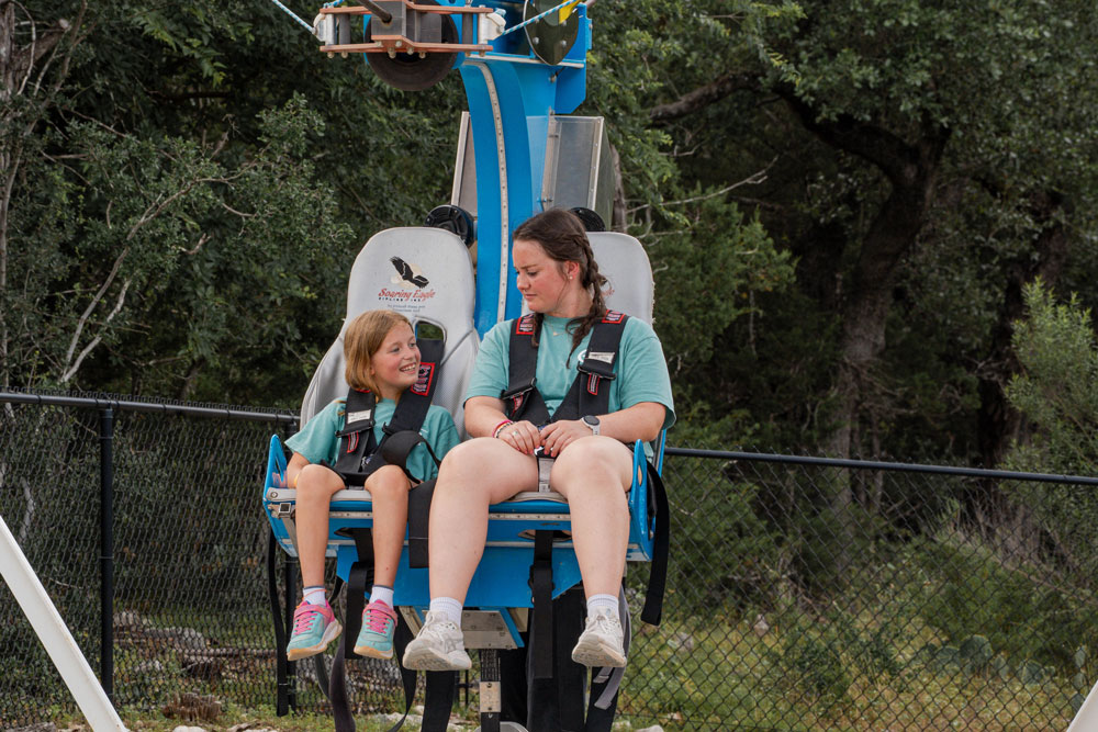 Two riders in teal shirts sit strapped into a blue amusement ride chair, awaiting departure with safety harnesses secured.