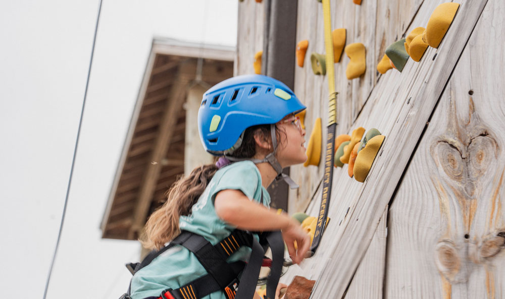 Child wearing a blue climbing helmet and harness climbs a wooden outdoor rock wall with yellow holds.